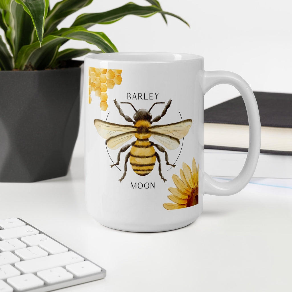 Barley Moon Botanicals 15 oz white ceramic mug featuring a bee, honeycomb, and flower illustration on a desk.