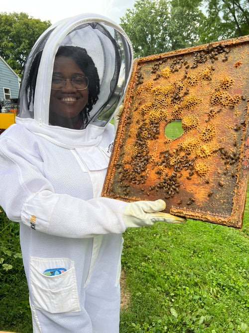 A smiling woman in a white protective beekeeping suit holding a wooden honeycomb frame at Barley Moon Botanicals.