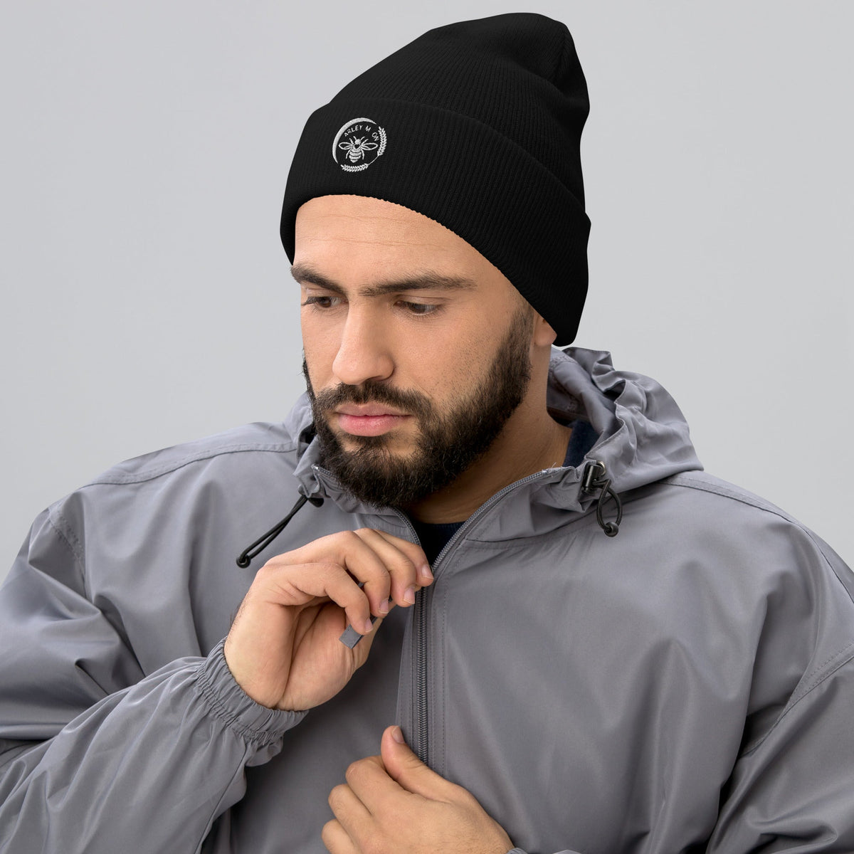 Bearded man wearing a black Barley Moon Botanicals cuffed beanie with a white bee and moon logo on a grey background