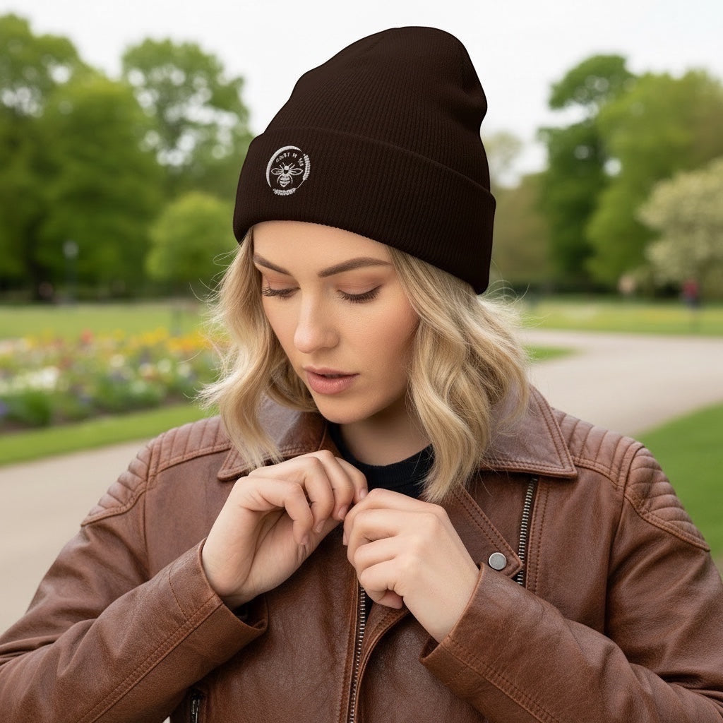 Woman wearing a brown Barley Moon Botanicals cuffed beanie with a white bee and moon logo in a park setting.