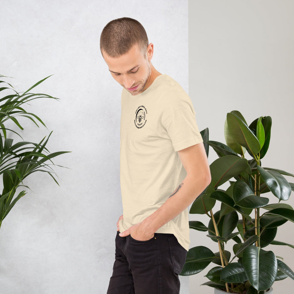 Man wearing a cream Barley Moon Botanicals unisex t-shirt with a small bee logo, standing between two indoor plants.