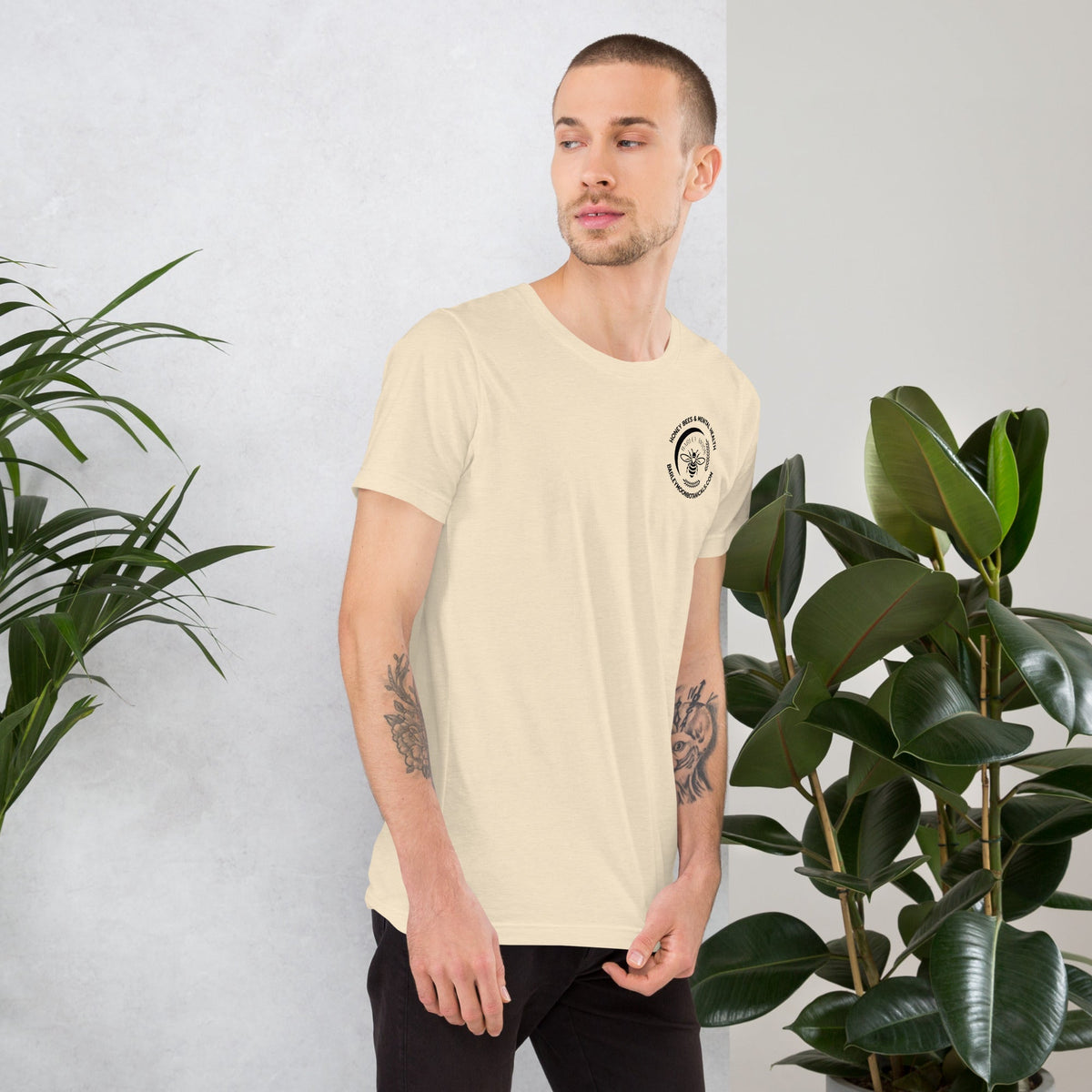 A man wearing a Barley Moon Botanicals cream unisex t-shirt with a small bee logo, standing by indoor plants.