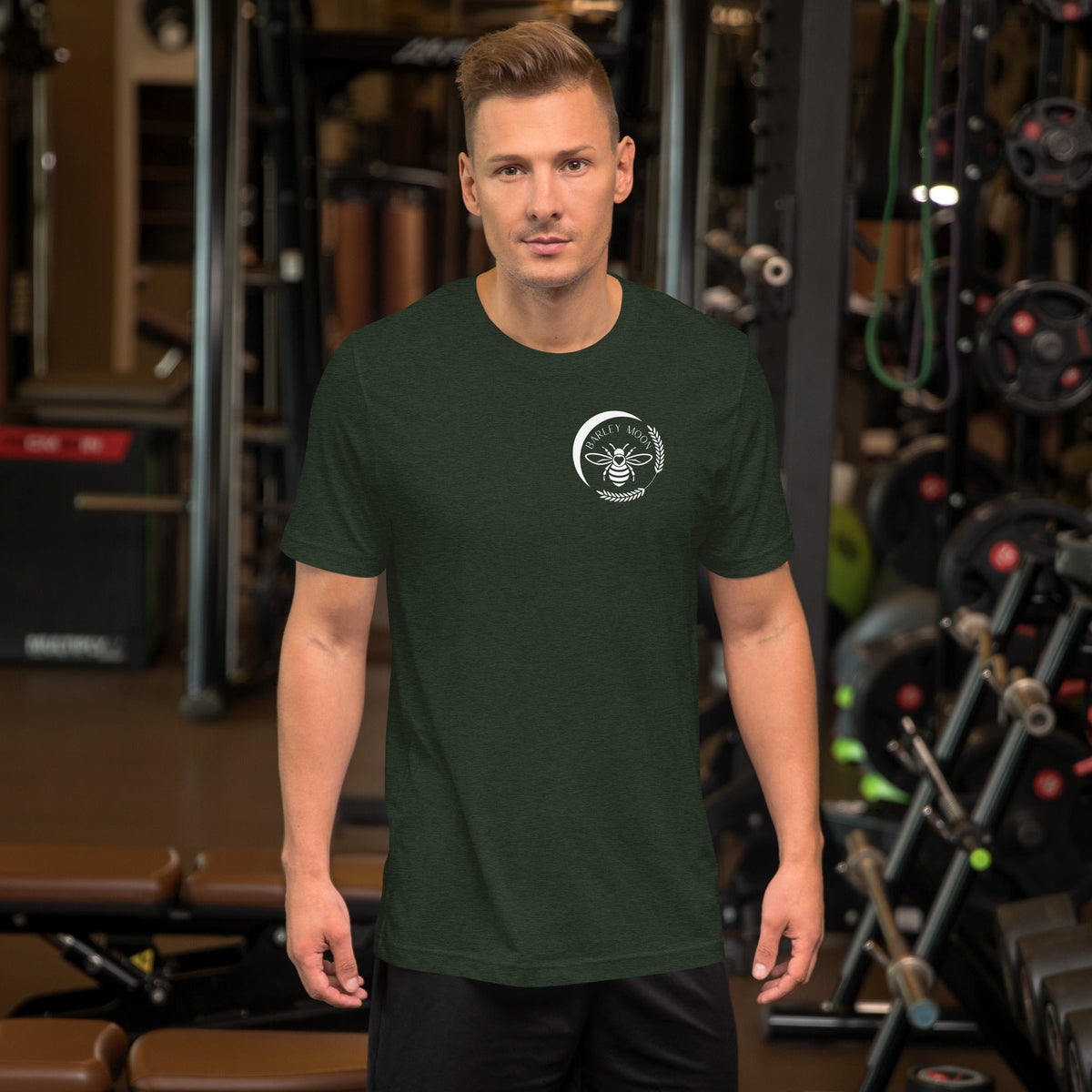 A man wearing a dark green Barley Moon Botanicals t-shirt with a white bee and crescent moon logo in a gym.