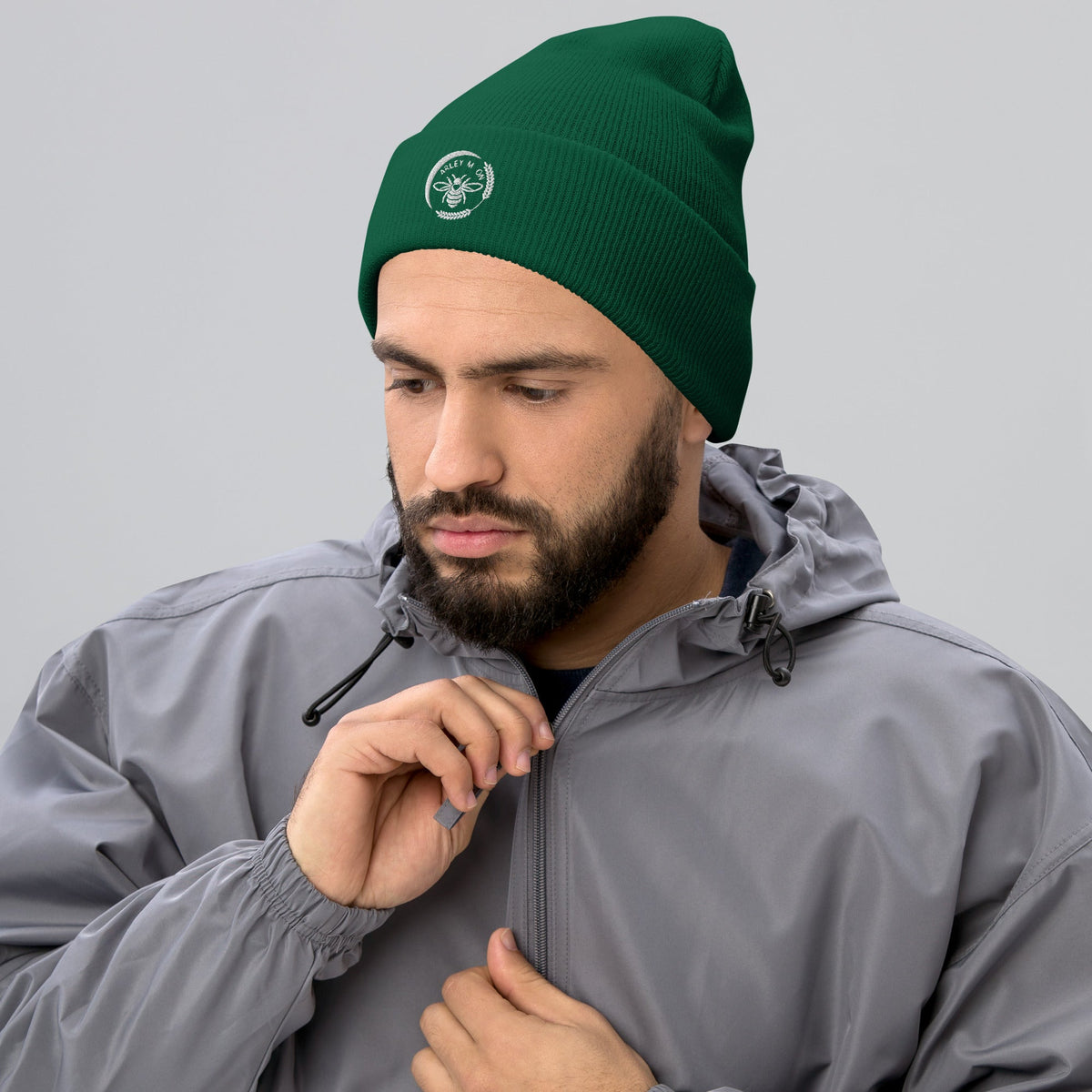 A bearded man wearing a green Barley Moon Botanicals cuffed beanie with a white bee and moon logo on a grey background.