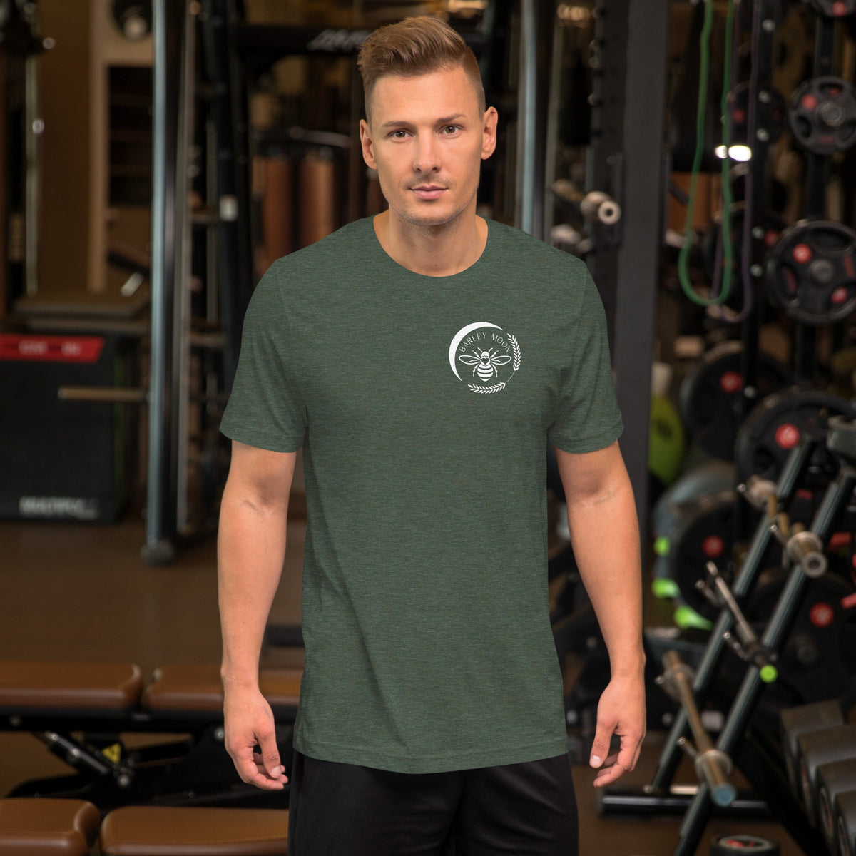Man wearing a Barley Moon Botanicals olive green unisex t-shirt with a white bee logo in a gym setting.