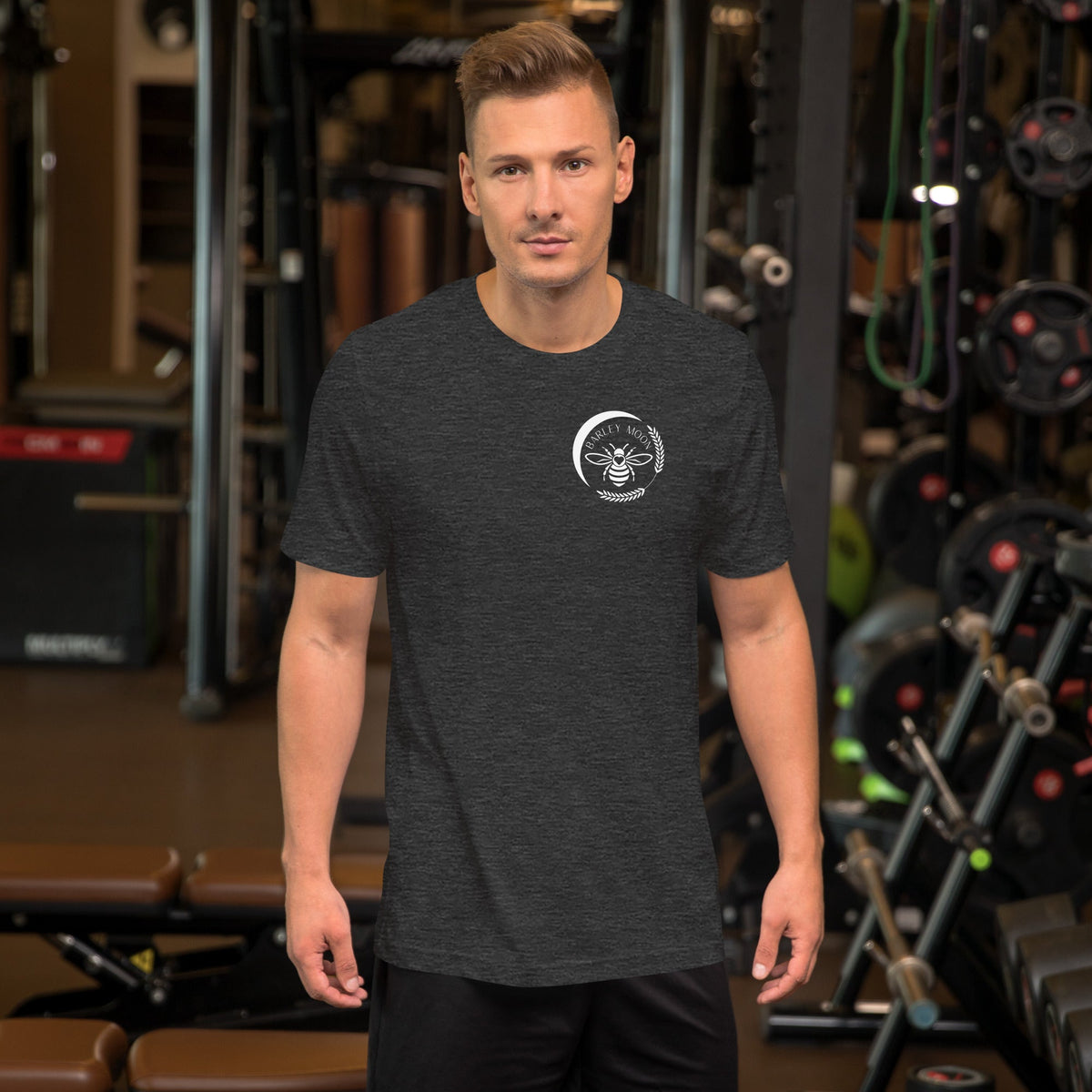 A man wearing a dark grey Barley Moon Botanicals t-shirt with a white bee and crescent moon logo in a gym setting.
