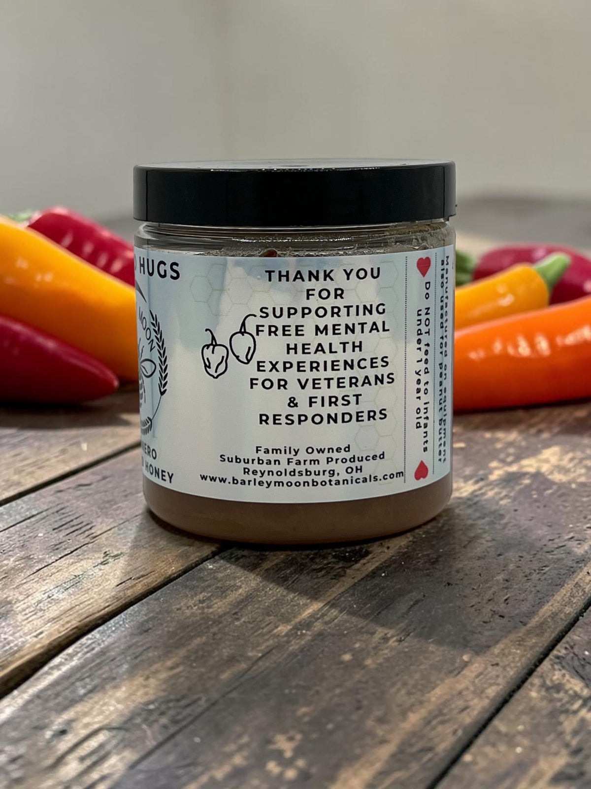 Barley Moon Botanicals Heated Hugs Habanero Honey jar on a wooden table with fresh peppers in the background.