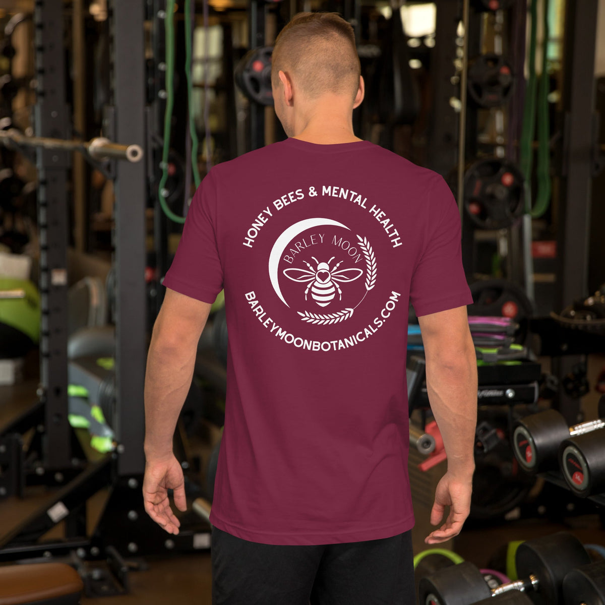 A man wearing a maroon Barley Moon Botanicals t-shirt with a white bee and crescent moon logo in a gym setting.