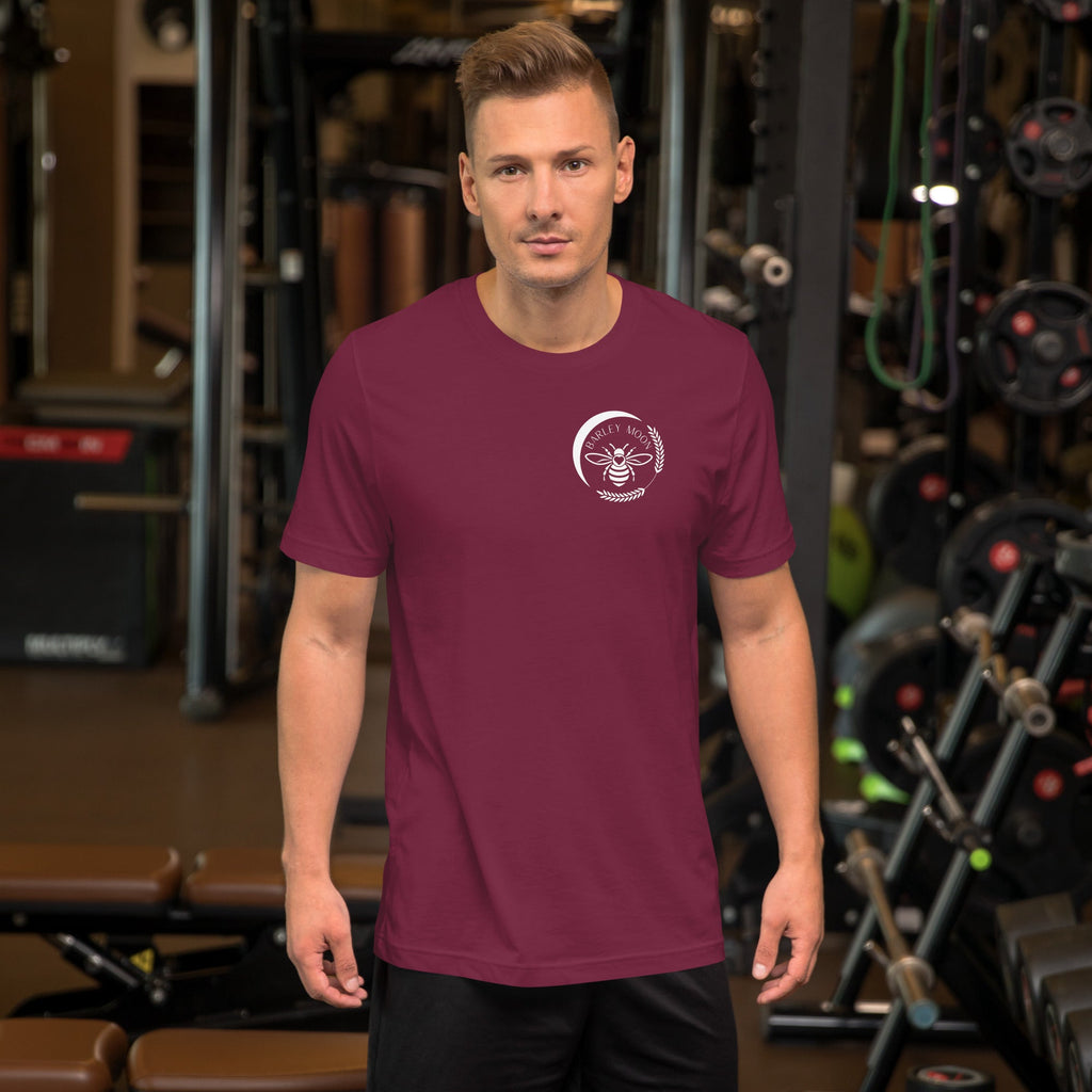 A man wearing a maroon Barley Moon Botanicals unisex t-shirt with a white bee logo on the chest in a gym setting.
