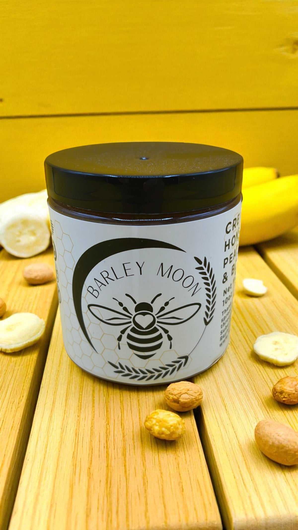 Barley Moon Botanicals Nutty Nana peanut butter and banana creamed honey jar on a wooden surface with fresh fruit