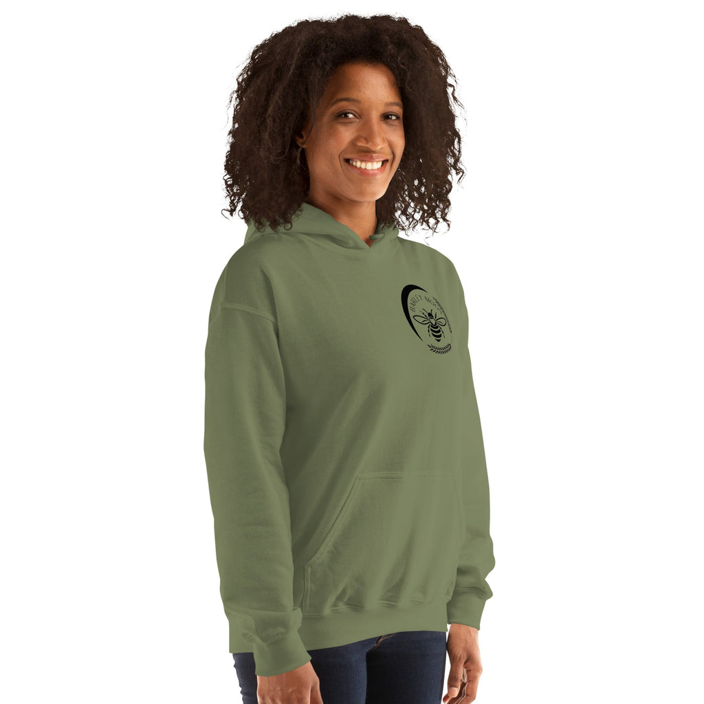 A woman wearing a sage green Barley Moon Botanicals unisex hoodie featuring a black bee and crescent moon logo.