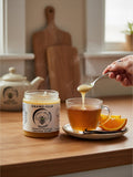 Barley Moon Botanicals Orang-illa orange vanilla creamed honey jar next to a steaming cup of tea with orange slices.
