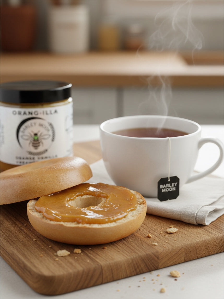 Barley Moon Botanicals Orang-illa creamed honey spread on a bagel next to a steaming cup of tea on a wooden board.