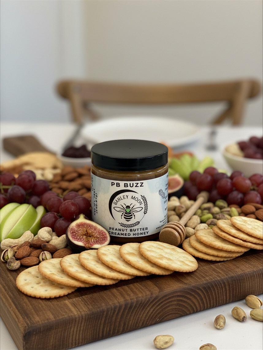 Barley Moon Botanicals PB Buzz peanut butter creamed honey jar on a wooden charcuterie board with fruit and crackers.