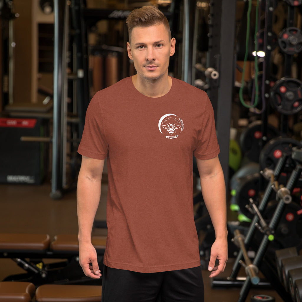 Man wearing a rust-colored Barley Moon Botanicals unisex cotton t-shirt with a white bee logo in a gym setting.