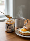 Barley Moon Botanicals Smoke-apeno smoked jalapeño creamed honey jar with a spoon, bagel, and steaming mug on a table