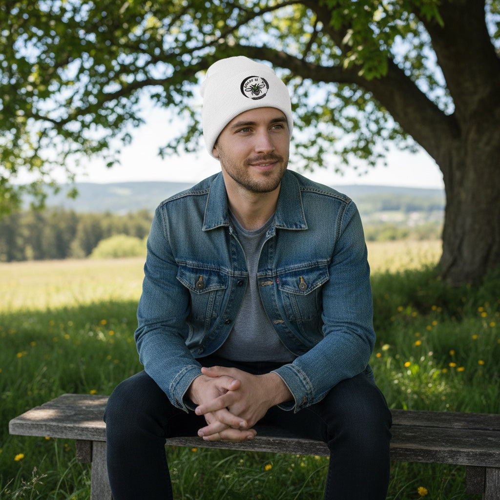 Man wearing a white Barley Moon Botanicals cuffed beanie with a black bee and moon logo while sitting on a bench.